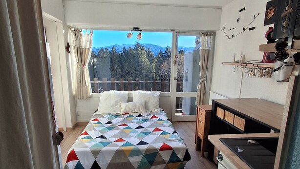 Une chambre avec un grand lit, une petite kitchenette et un balcon offrant une vue sur les arbres et les montagnes.