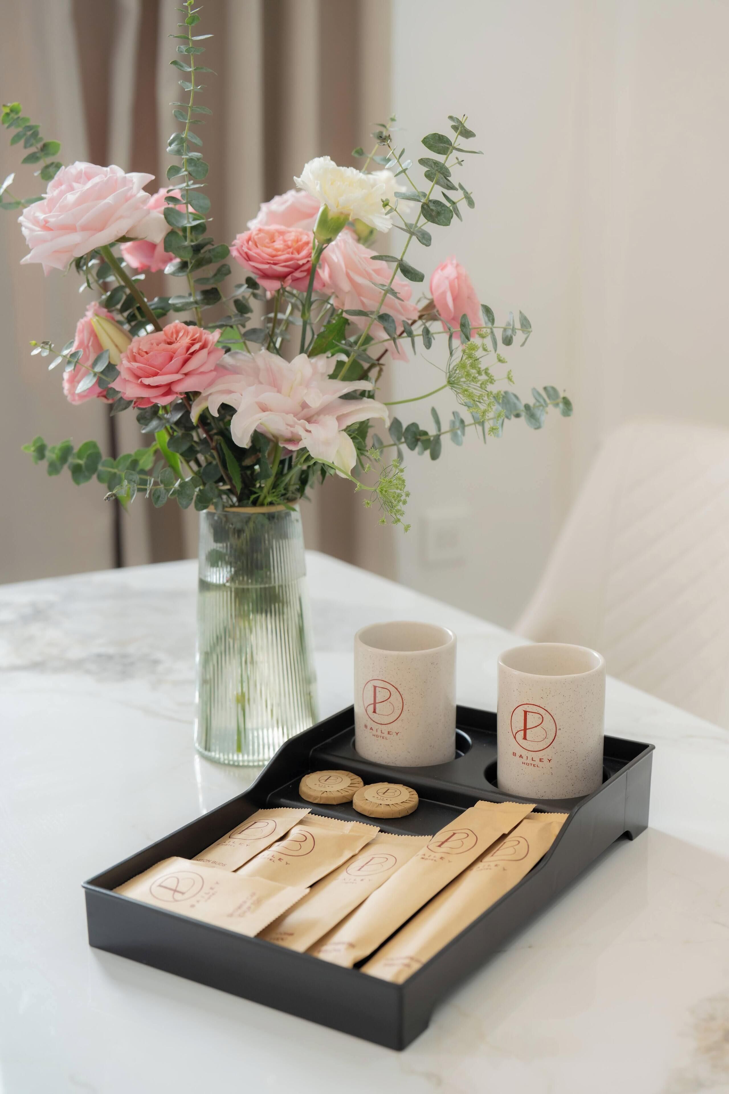 A table with a vase of pink and white flowers, two mugs with a logo, and a tray with tea bags.
