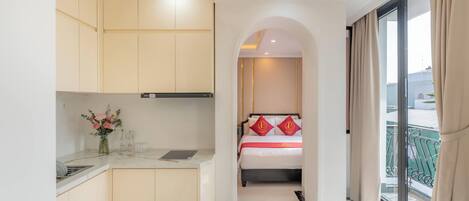 A hotel room with a kitchenette, a bed with red pillows, and a balcony with curtains.