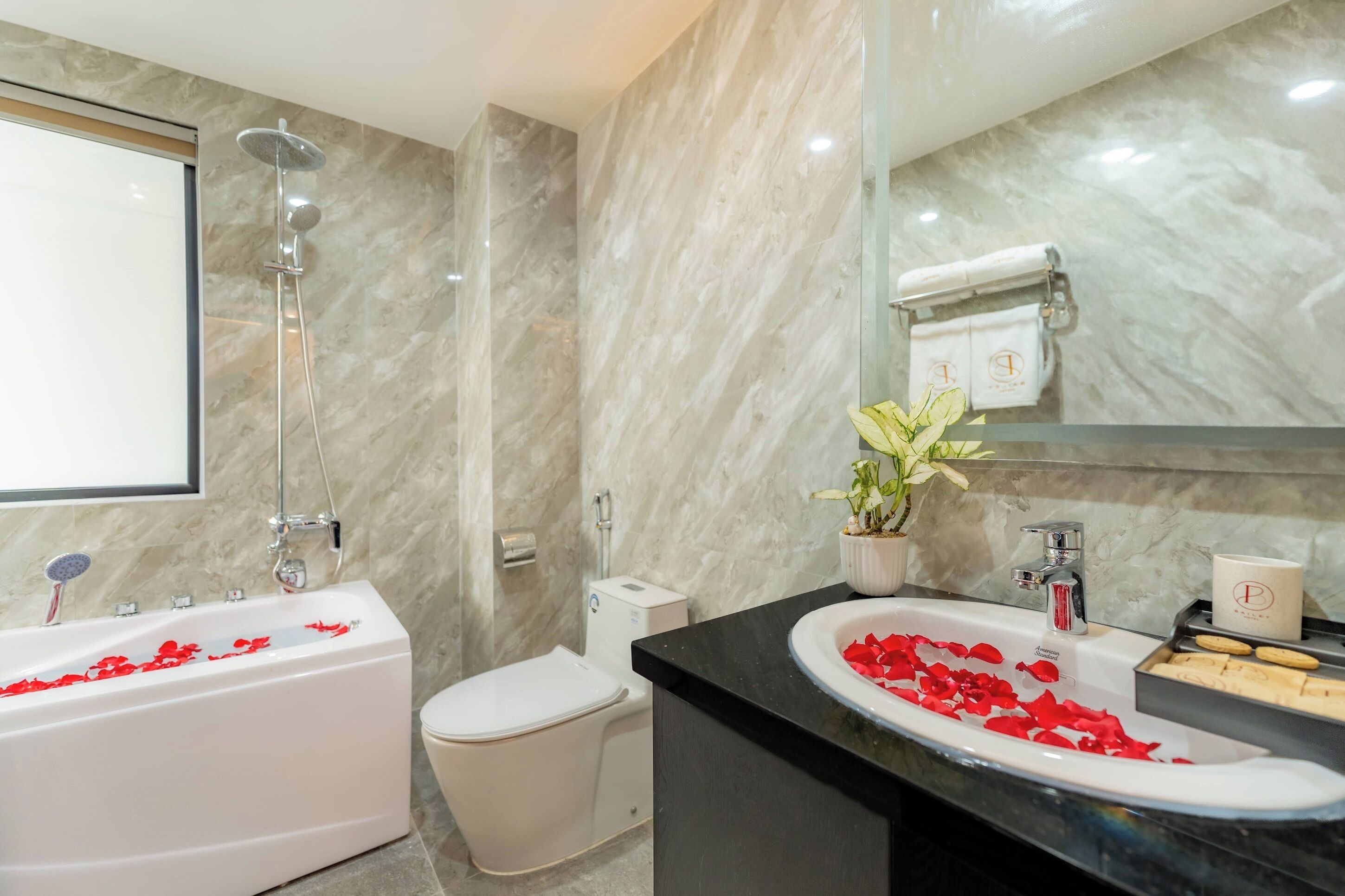 Un baño moderno con pared de mármol, una bañera con pétalos de rosa, un inodoro, un lavabo con espejo y una planta en maceta.