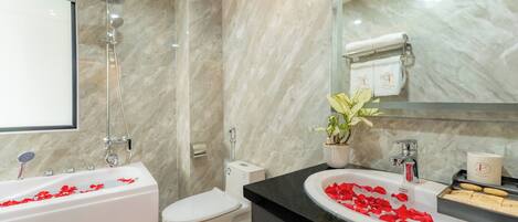 Un baño moderno con pared de mármol, una bañera con pétalos de rosa, un inodoro, un lavabo con espejo y una planta en maceta.