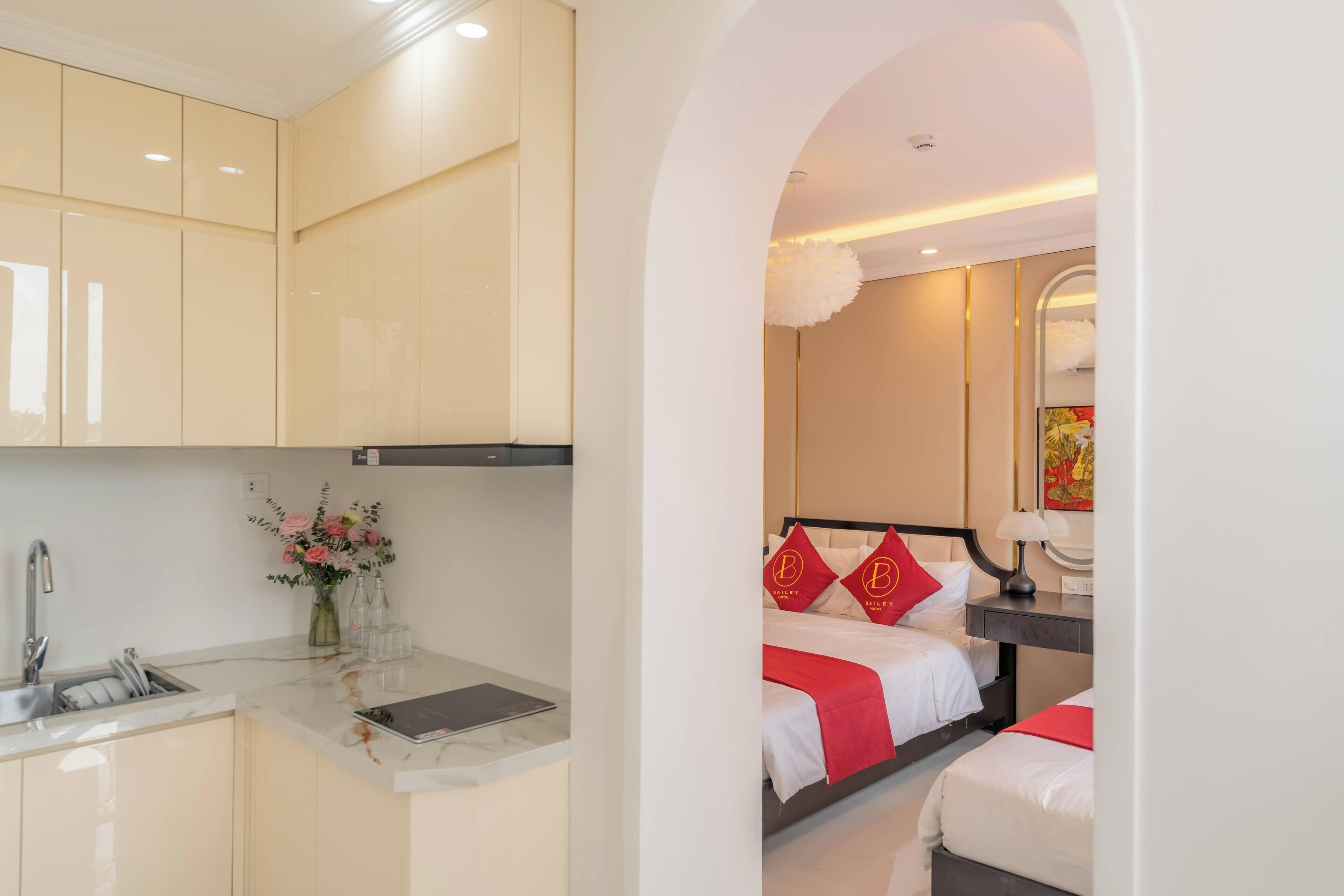 A modern hotel room with a kitchen area, a bed with red and white bedding, and a small desk with a lamp.