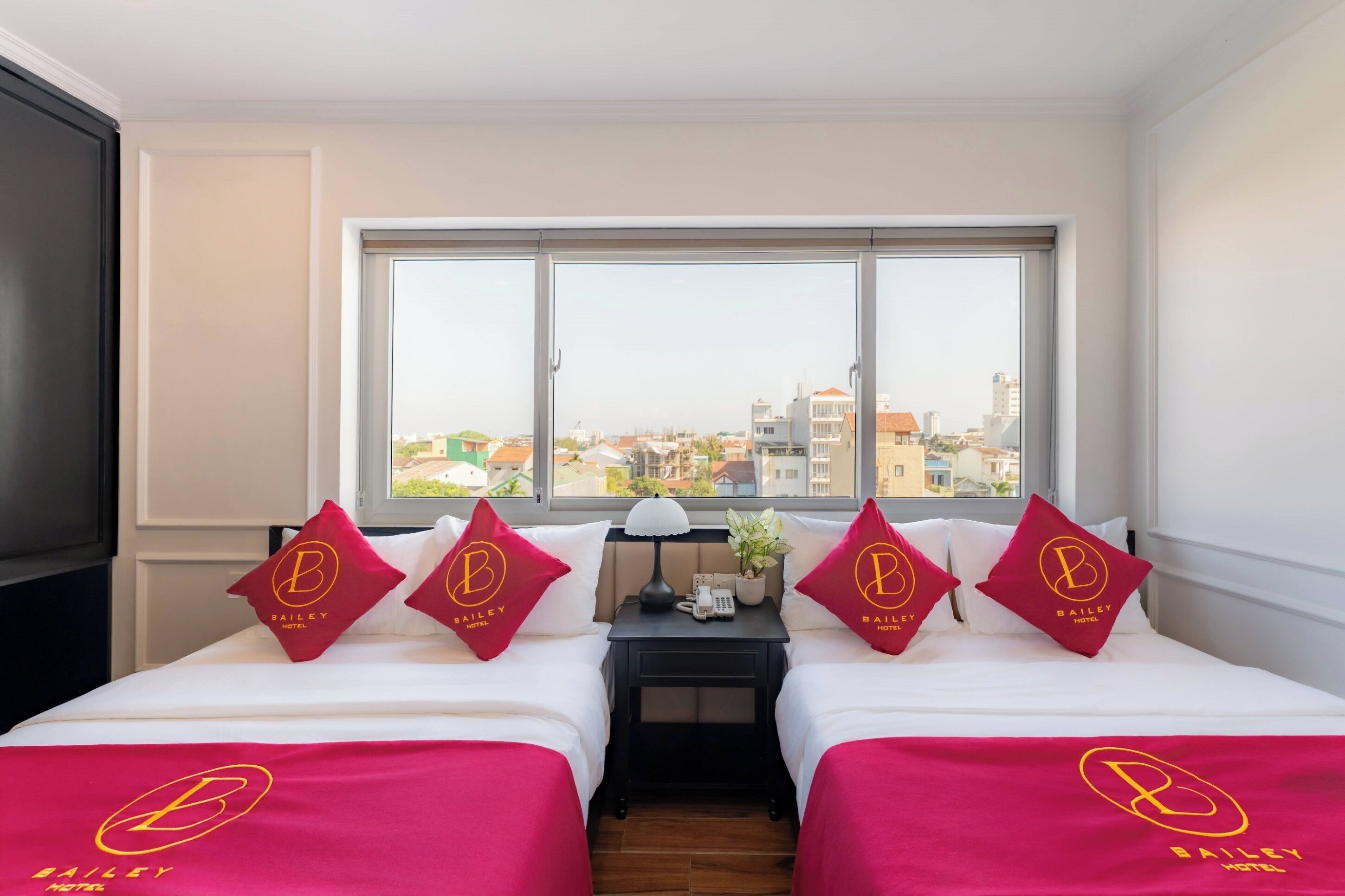 Ein Hotelzimmer mit zwei Betten, jedes mit einem Kopfkissen mit dem Bailey Hotel Logo, einem Nachttisch mit Lampe und Telefon sowie einem großen Fenster mit Blick auf die Stadt.