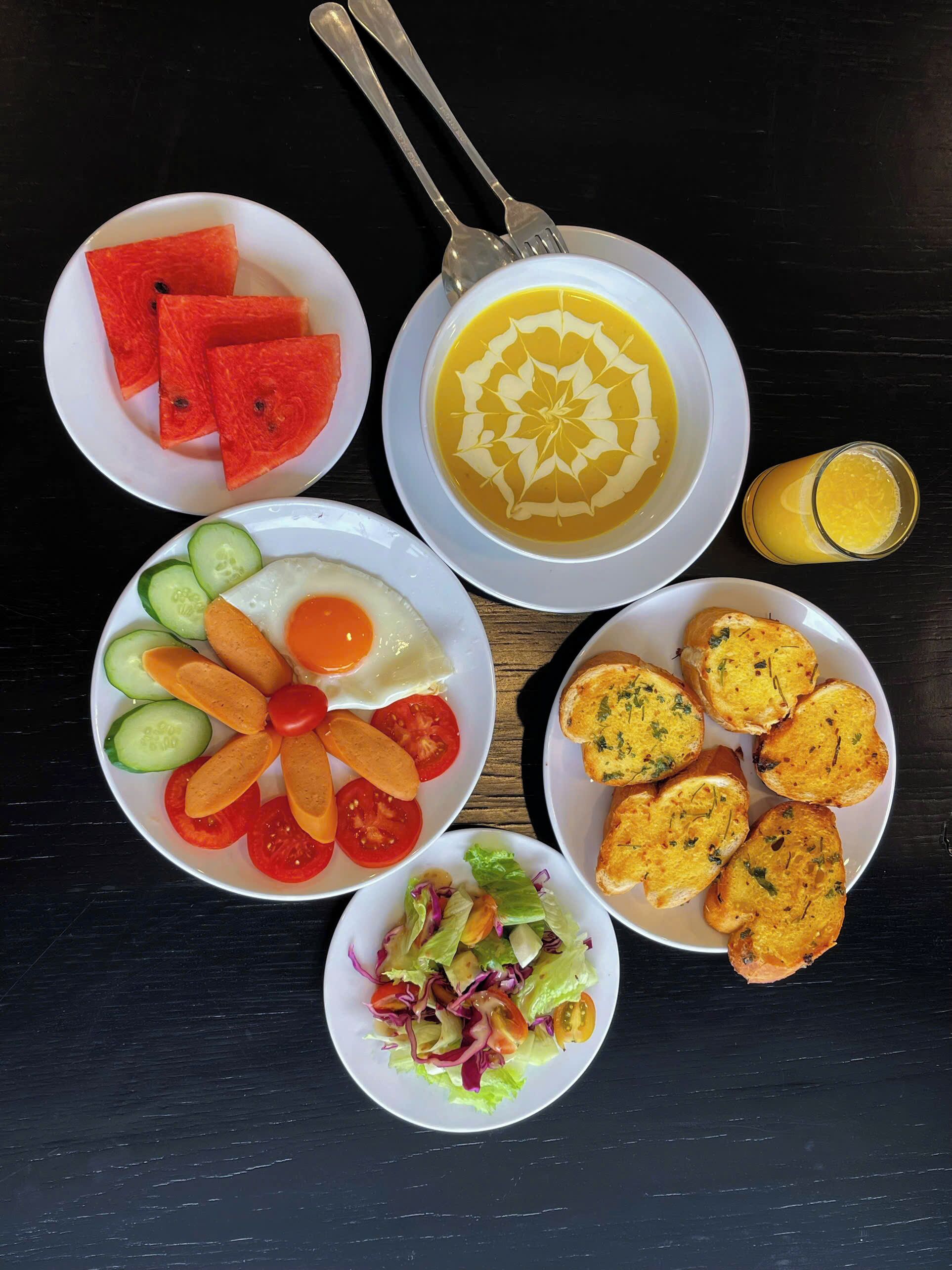 Een tafel met verschillende gerechten, waaronder stukken watermeloen, soep, eieren, plakjes komkommer, tomaten, een salade en knoflookbrood.