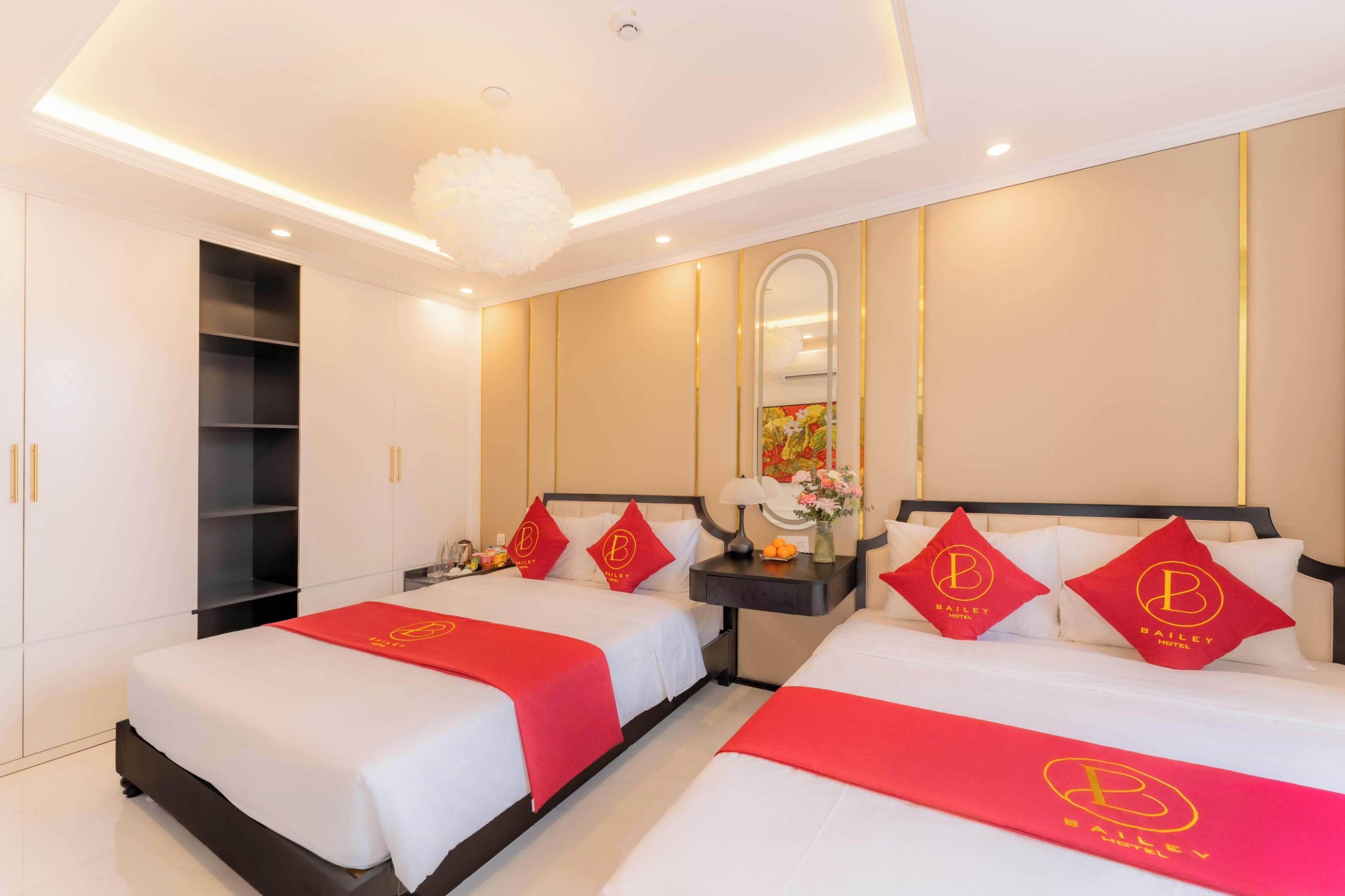 A hotel room with two beds, each with a red bedspread and pillows featuring a logo. There is a nightstand with a lamp and a vase of flowers. The room has a modern design with a large mirror and a ceiling light fixture.