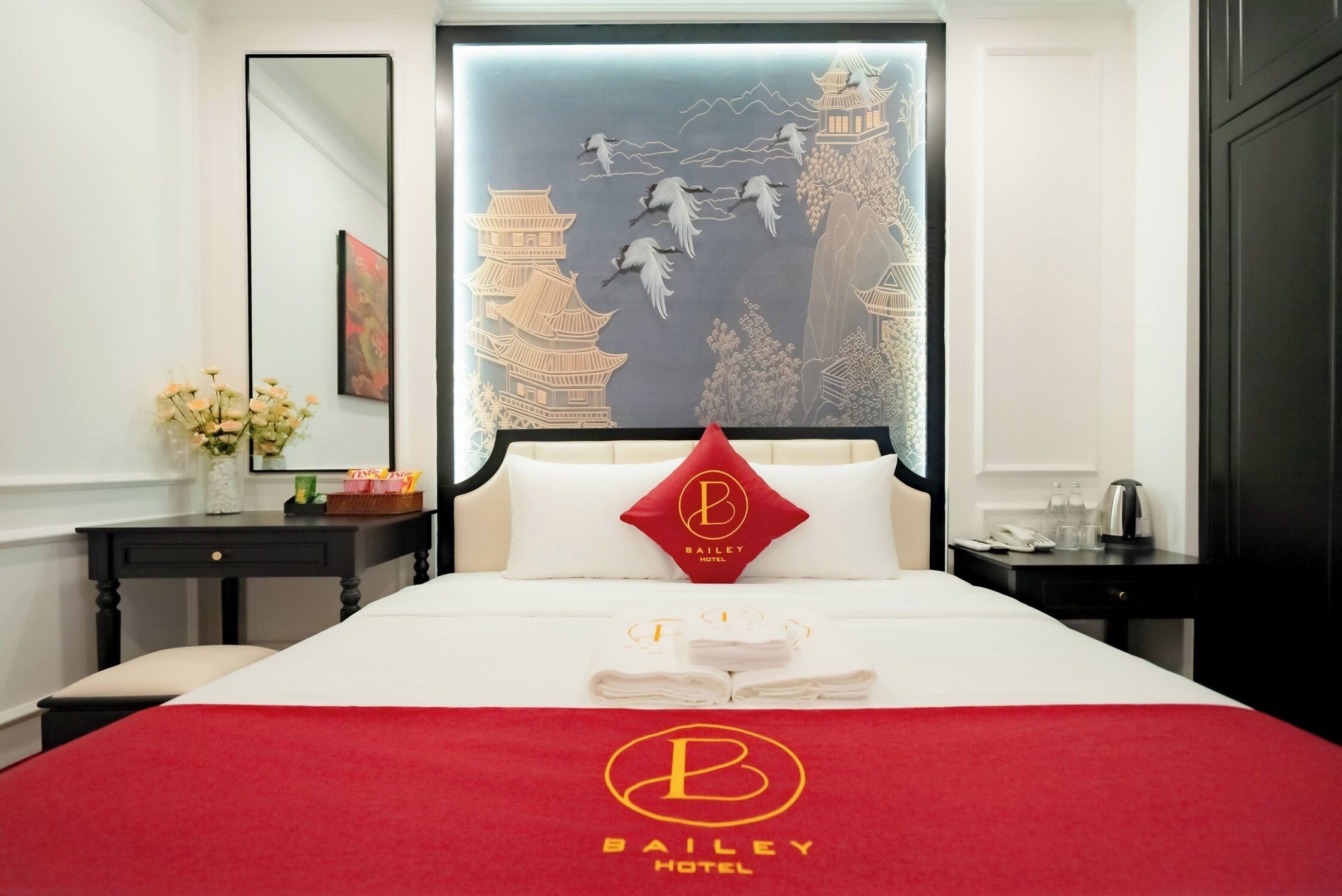 A hotel room with a large bed, a red throw blanket, a pillow with the hotel logo, a nightstand with a lamp, and a wall mural.