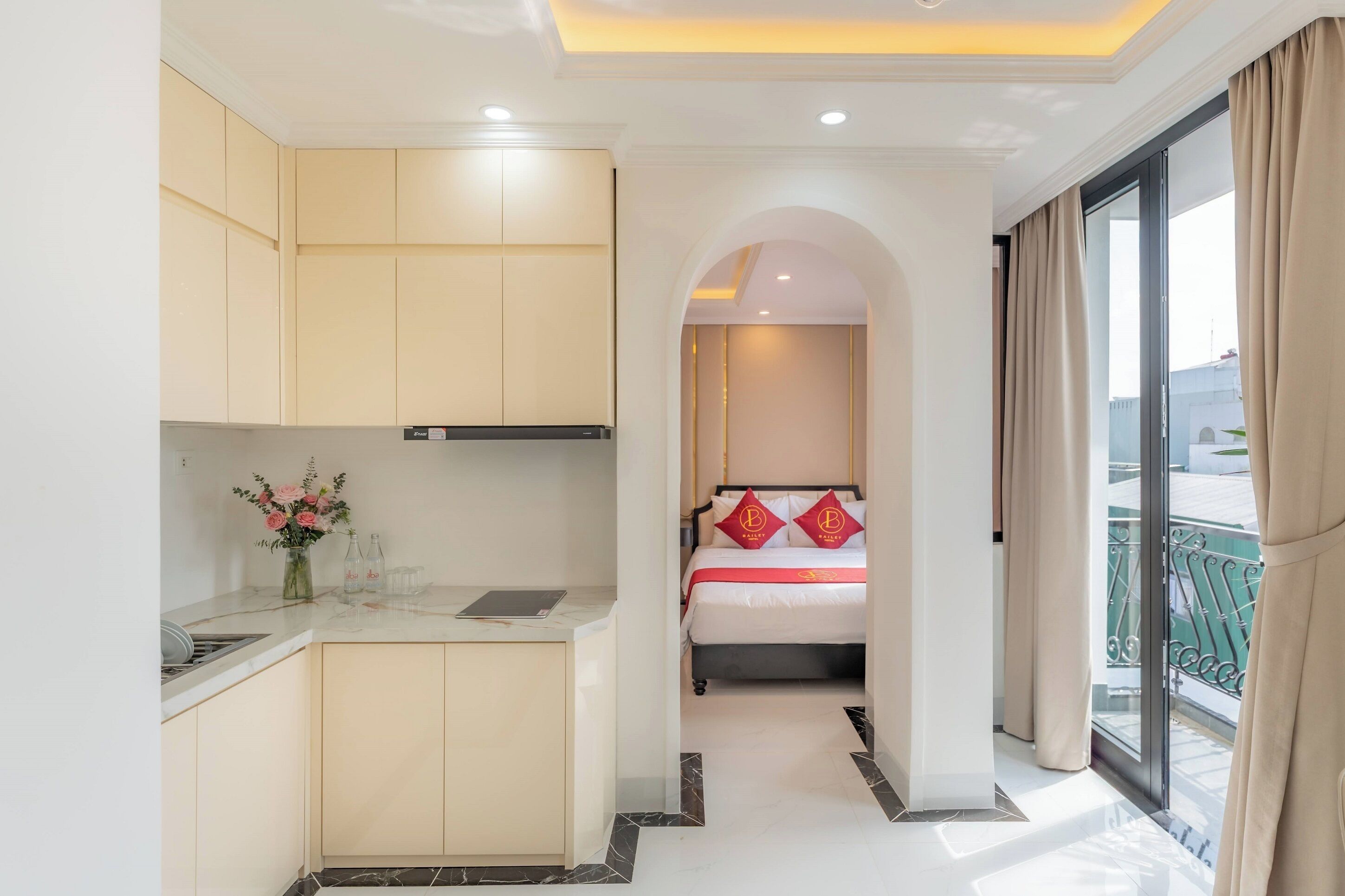 Una habitación de hotel con kitchenette, una cama con almohadas rojas y un balcón con cortinas.