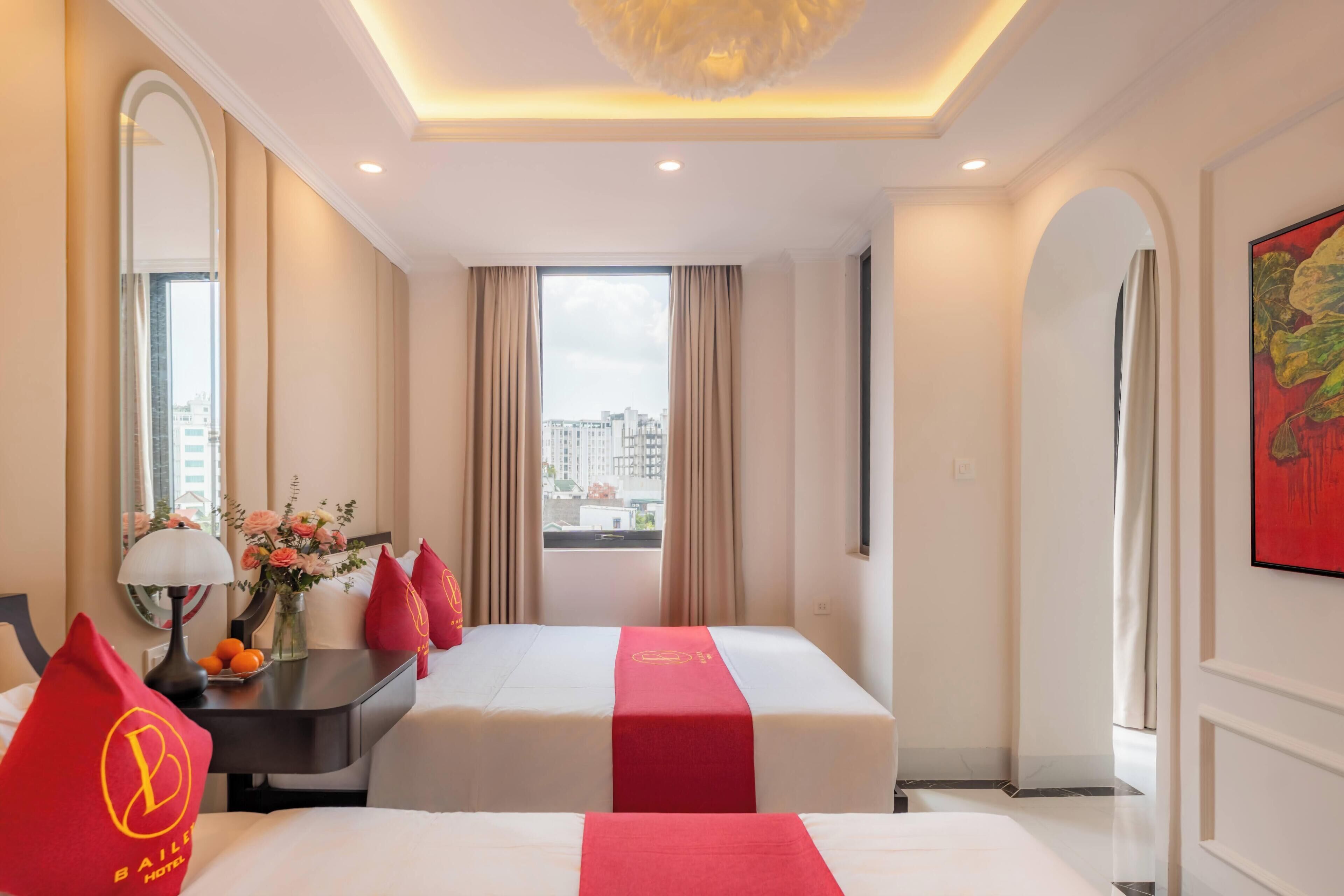 A hotel room with two beds, a desk with a lamp, a vase with flowers, and a city view through the window.