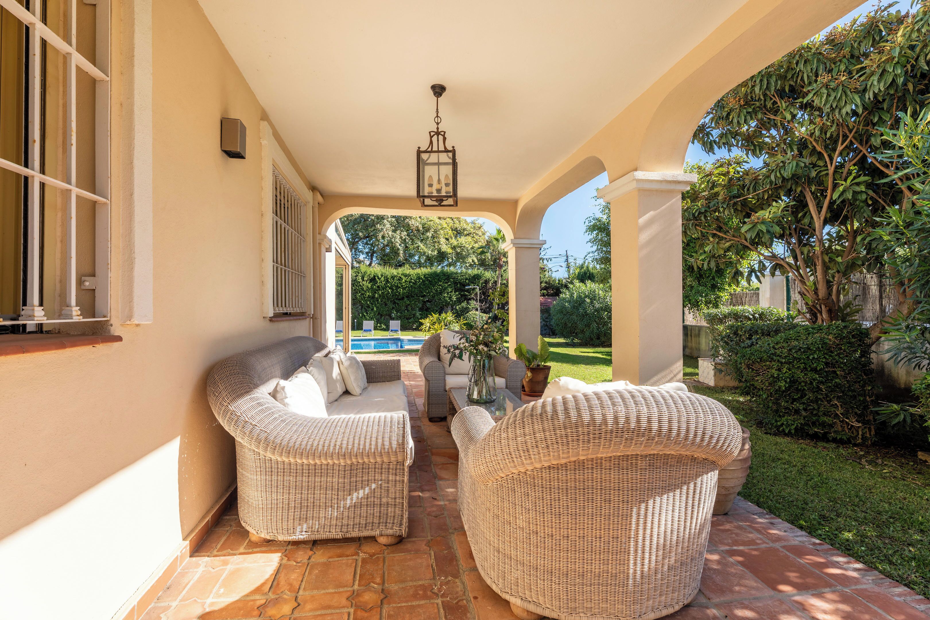 A covered patio with wicker furniture, a pool in the background, and a well-maintained garden.