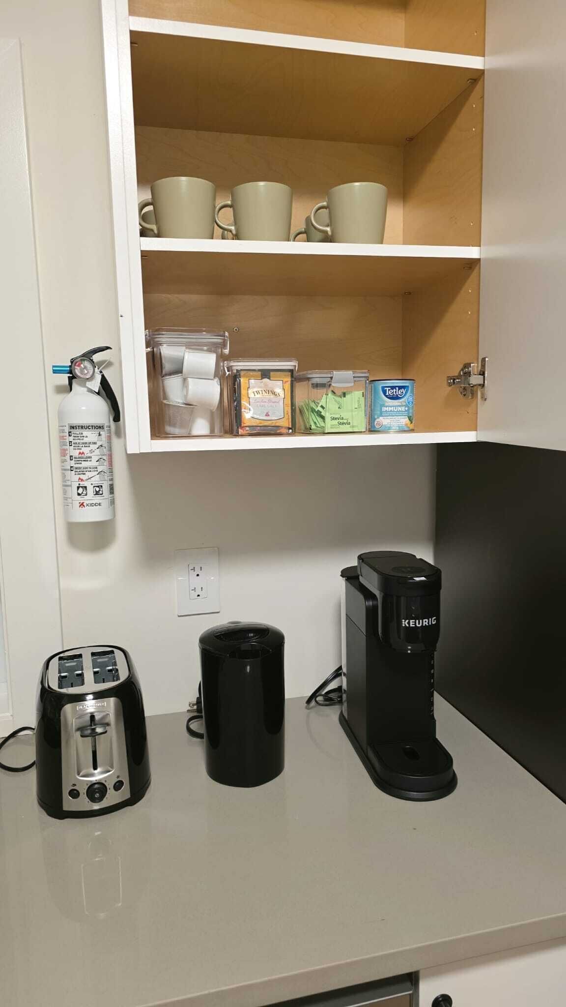 A kitchenette with a coffee maker, toaster, trash can, and a cabinet containing cups and food items.