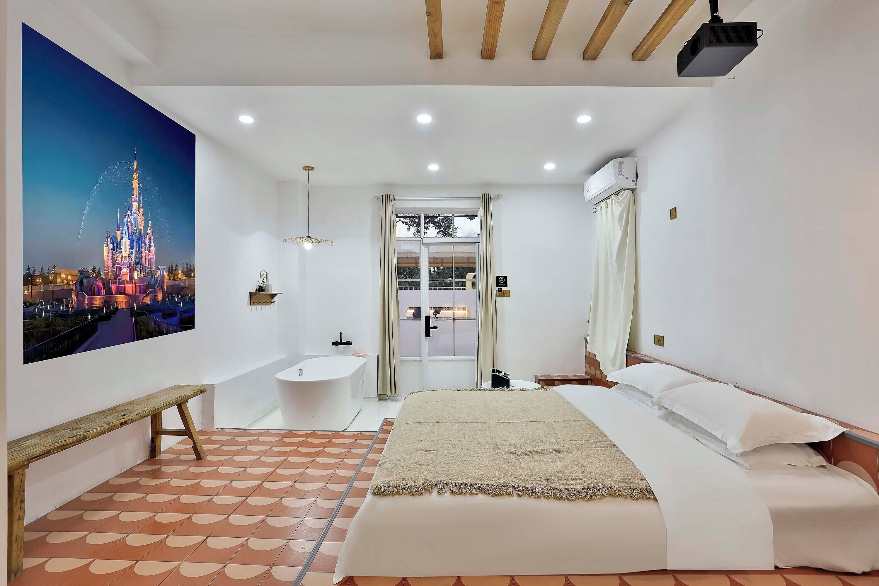 Habitación de hotel con un mural grande de un castillo, un banco de madera, una cama con ropa blanca, un baño con bañera y un proyector en el techo.