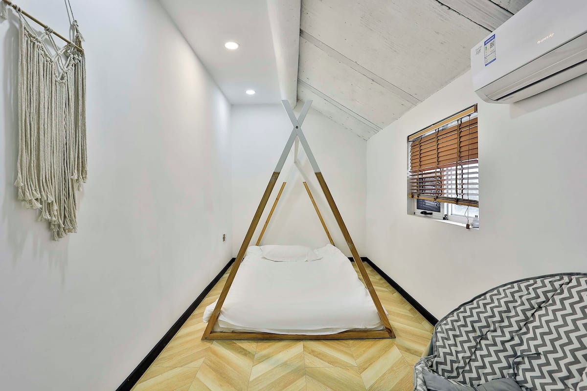 Une chambre avec un lit, une décoration murale en macramé, une fenêtre avec des stores et un appareil de climatisation.
