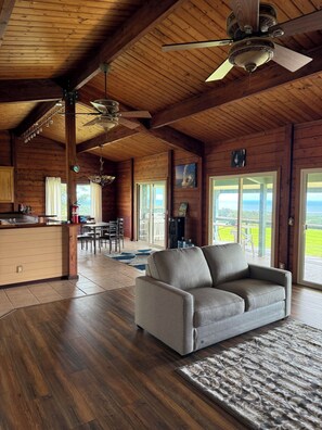 Amplia sala de estar con sofá, pisos de madera, ventiladores de techo y vistas a través de grandes ventanales.