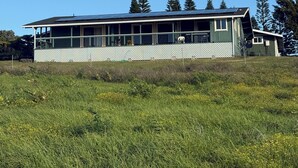 A single-story house with a large deck, surrounded by a white picket fence, set in a grassy field with trees in the background.