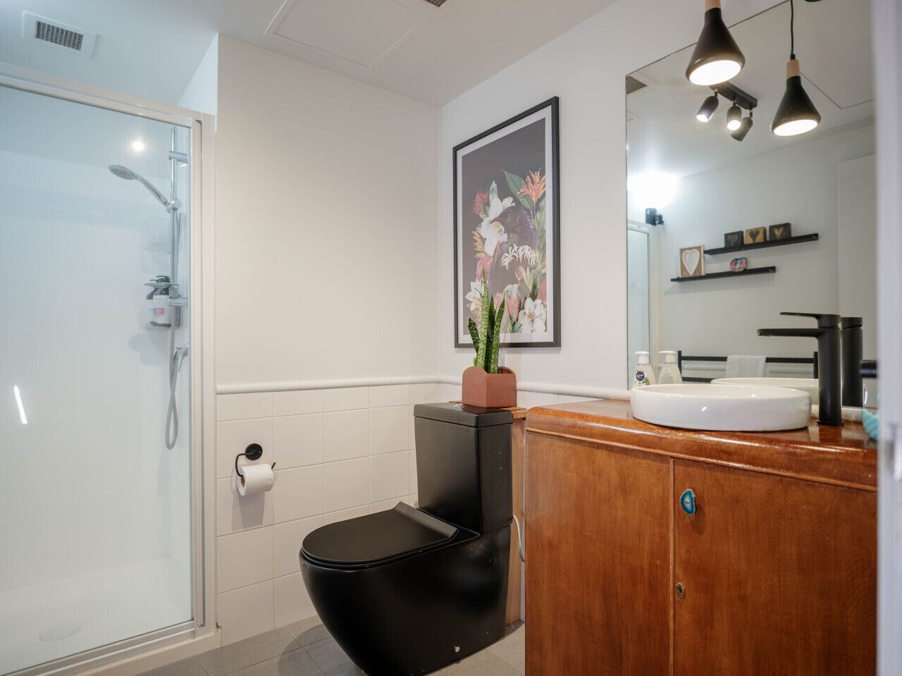 A modern bathroom with a glass shower enclosure, a black toilet, a wooden vanity with a white sink, and a framed floral artwork on the wall.