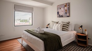 A modern bedroom with a large bed, a window with blinds, a bedside table with a lamp, and a painting on the wall. - The Paloma by Homello | Quiet • Central • Perfect for Long Stays (Auckland)