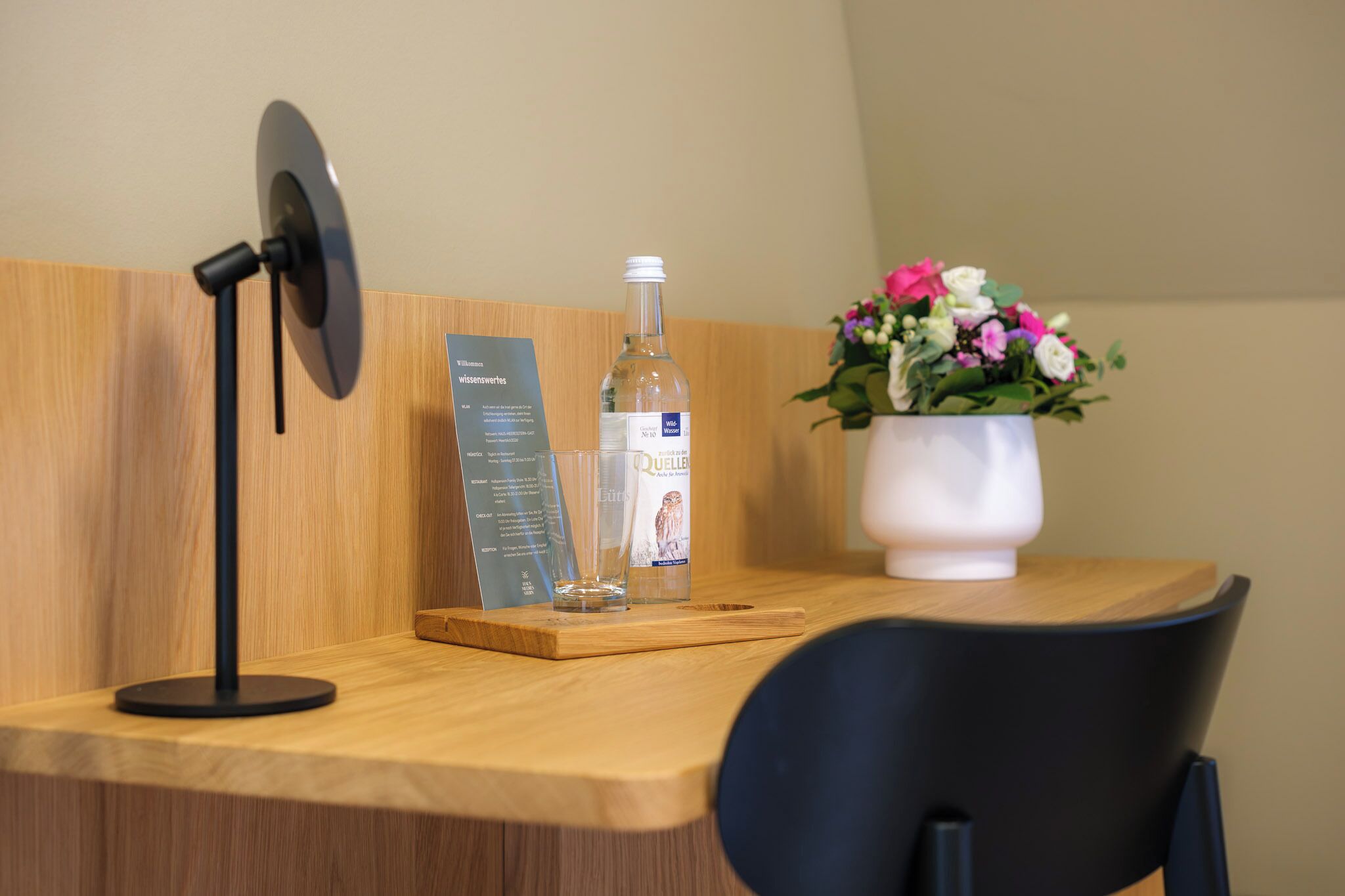 A wooden desk with a fan, a bottle of water, a glass, and a vase with flowers.