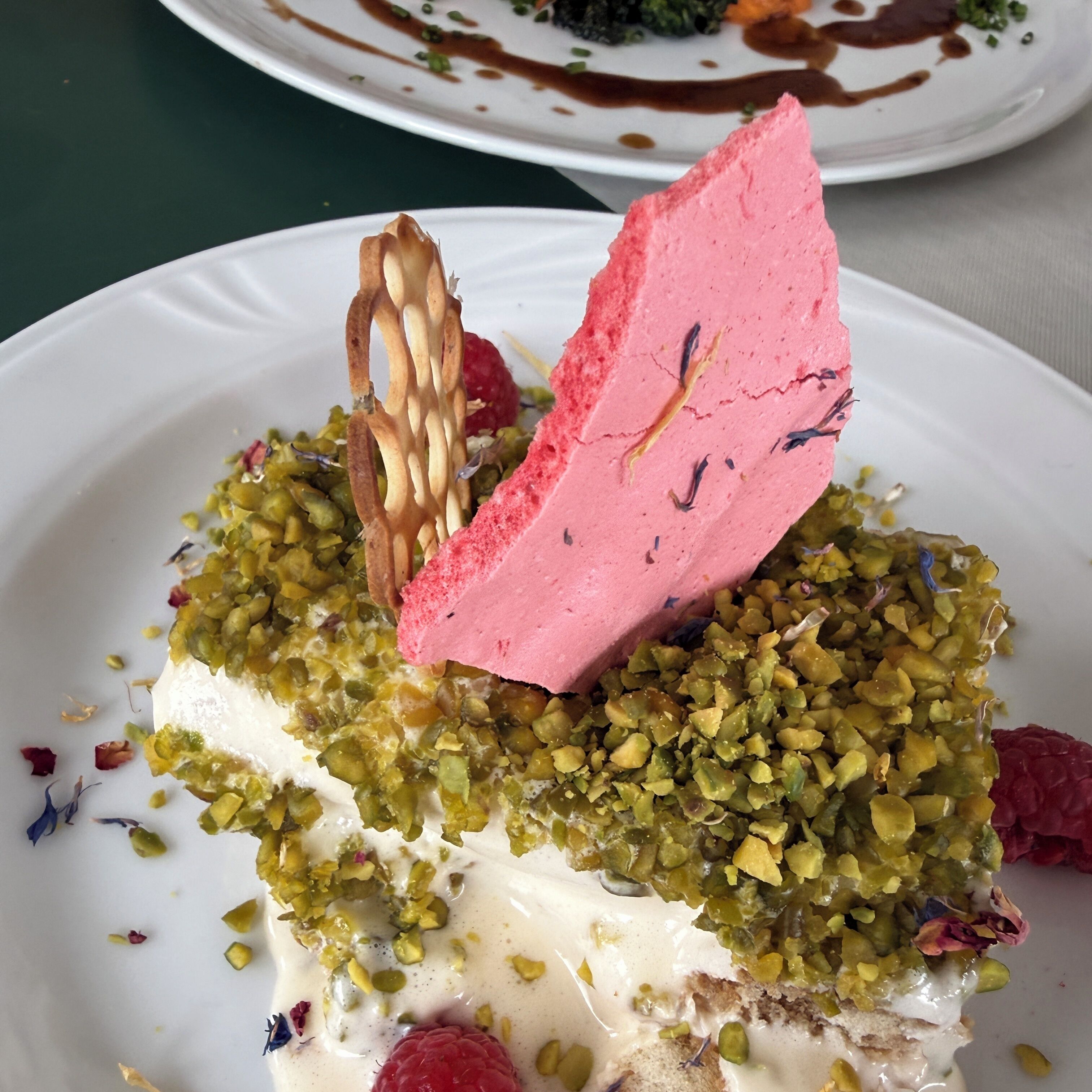 Un dessert aux pistaches, agrémenté d’un élément rose et d’un morceau de chocolat, présenté sur une assiette.