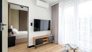 A modern hotel room with a wooden floor, a flat-screen TV mounted on the wall, a bedside table with a lamp, a bedside plant, and a large window with sheer curtains.