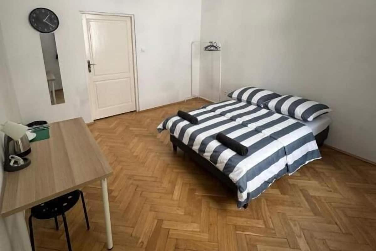 Una habitación pequeña y escasamente amueblada, con piso de madera en patrón de espiga, una cama con ropa de cama a rayas, un escritorio con una silla, una lámpara colgante y una puerta.