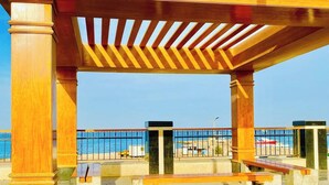 Puinen pergola, jossa on penkkejä, betonilattia ja metallikaide, jolta avautuu näkymä rannalle.