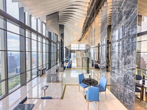 A spacious lobby with a high ceiling, large windows offering city views, a central staircase, and modern seating areas.