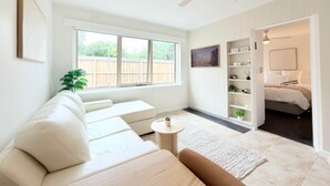En moderne stue med en hvid sofa, et lille rundt bord og en ventilator i loftet. Der er et stort vindue med udsigt til et træhegn udenfor.