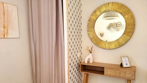 En del av ett rum med en beige gardin, en väggpanel med struktur, en rund spegel med guldfärgade ram, ett träbord med en vas och ett inramat certifikat, och en liten växt.