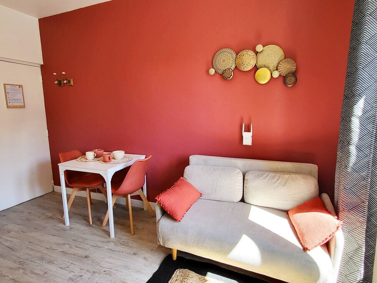 A room with a red wall, a white table with chairs, a grey sofa with red cushions, and a decorative wall piece.