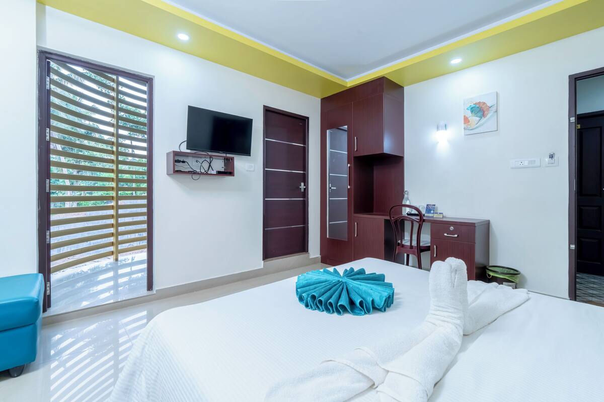 A hotel room with a bed, a TV mounted on the wall, a wardrobe, and a desk with a chair.