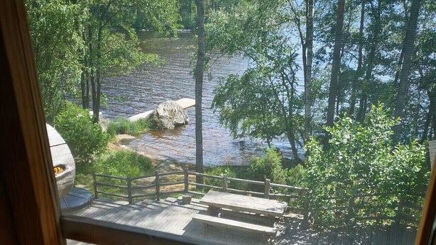 Une vue depuis une fenêtre donnant sur une terrasse en bois, avec une rivière et des arbres en arrière-plan.