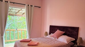 A bedroom with a bed, curtains, a nightstand, a lamp, and a view of greenery outside. - Villa Amarela - Itacimirim - Bahia - 5 suítes (Camaçari)
