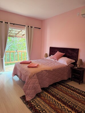 Een slaapkamer met een bed, gordijnen, een nachtkastje, een lamp en uitzicht op groen.