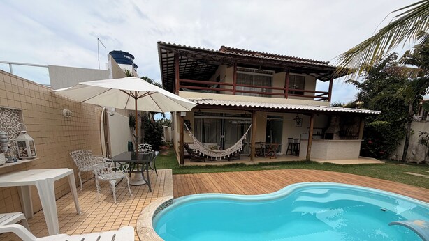 A backyard with a swimming pool, a dining area with chairs and a table, and a covered patio area. - Villa Amarela - Itacimirim - Bahia - 5 suítes (Camaçari)