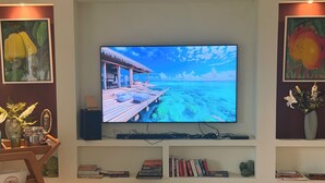 A flat-screen TV mounted on a wall with a wooden shelf below displaying books and decorative items. - Villa Amarela - Itacimirim - Bahia - 5 suítes (Camaçari)