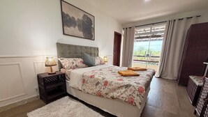 A bedroom with a bed, a nightstand, a lamp, a rug, and a window with curtains. - Villa Amarela - Itacimirim - Bahia - 5 suítes (Camaçari)