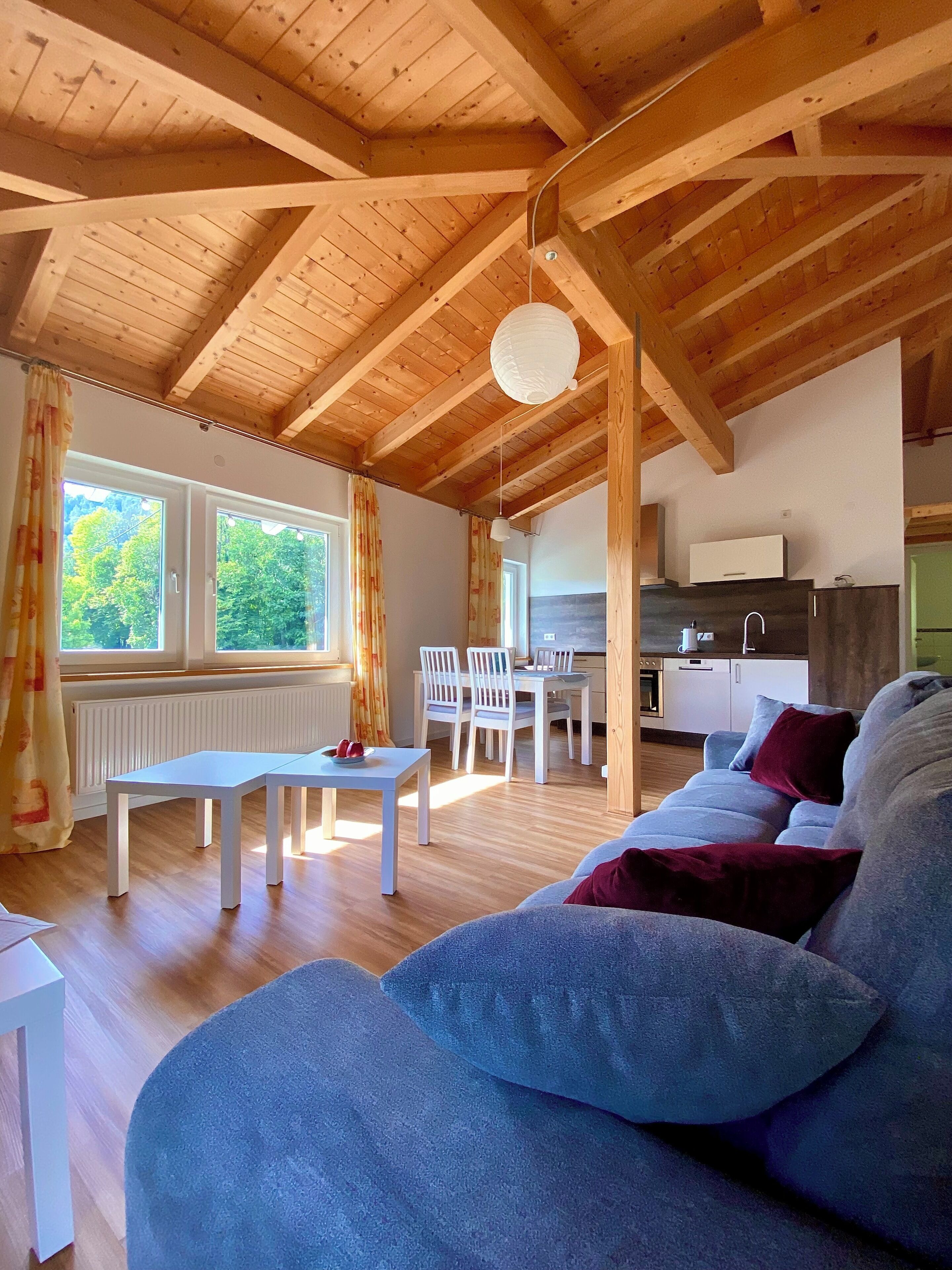 Une pièce spacieuse avec un haut plafond en bois, un coin repas avec des chaises blanches et une table, un coin cuisine et un espace détente avec un canapé et des coussins.