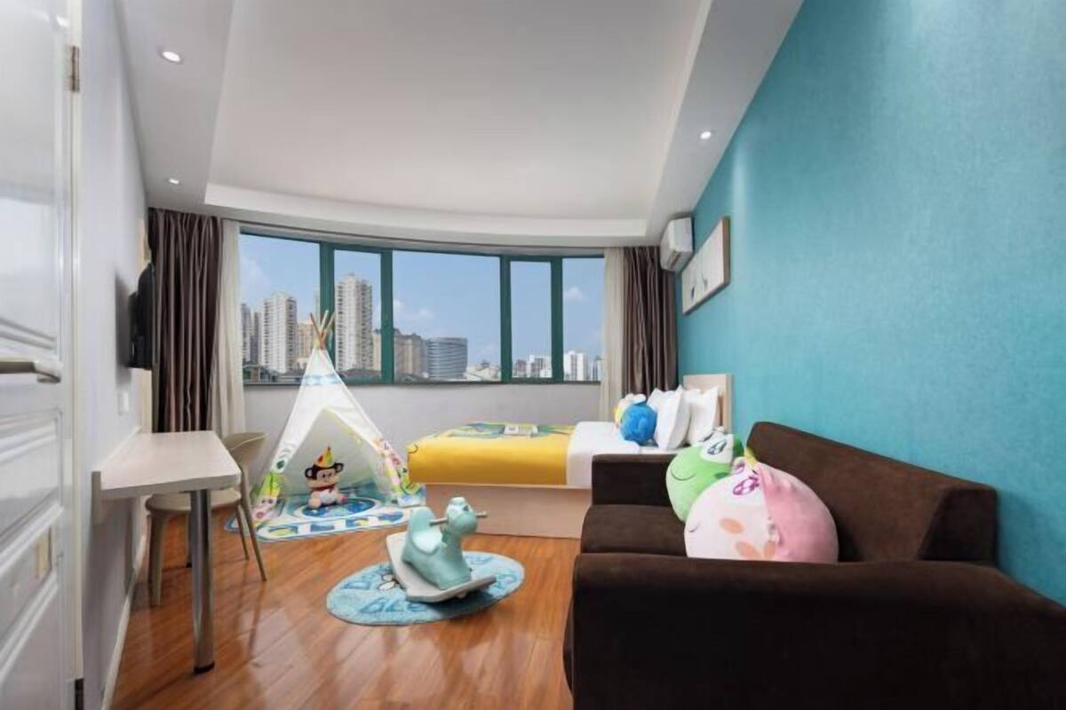 A modern hotel room with a large window, a bed, a sofa, a desk, and a child's play area.