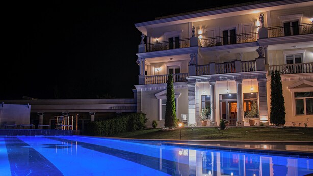 Una grande piscina ben illuminata, con finiture in marmo, circondata da un prato curato e un imponente edificio a più piani con balconi e dettagli architettonici in stile classico.
