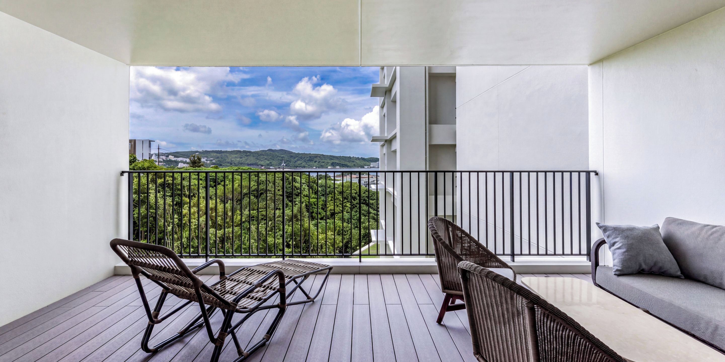 A balcony with wicker furniture, a view of greenery and a distant landscape, and a white building in the background.