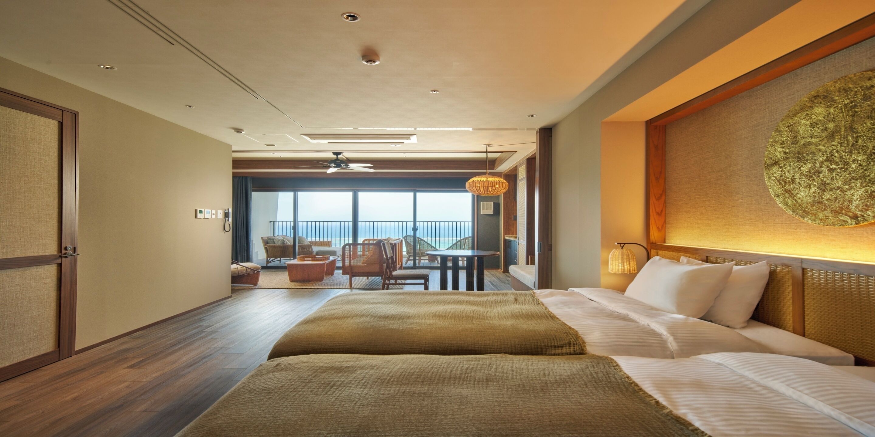 A hotel room with a bed, a dining area with chairs, a view of the sea, and a ceiling fan.