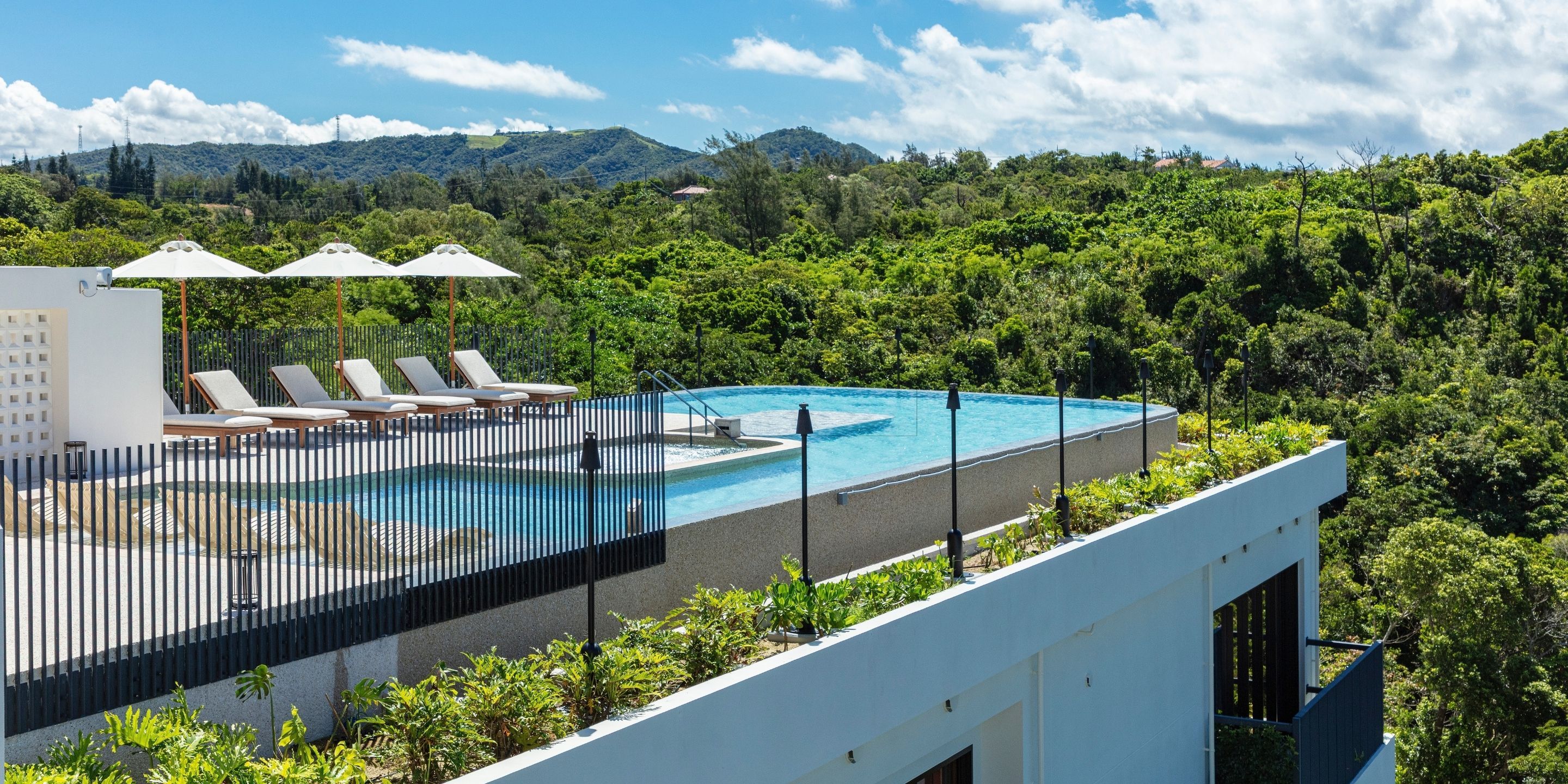 A rooftop pool with lounge chairs and umbrellas, surrounded by lush greenery and hills.