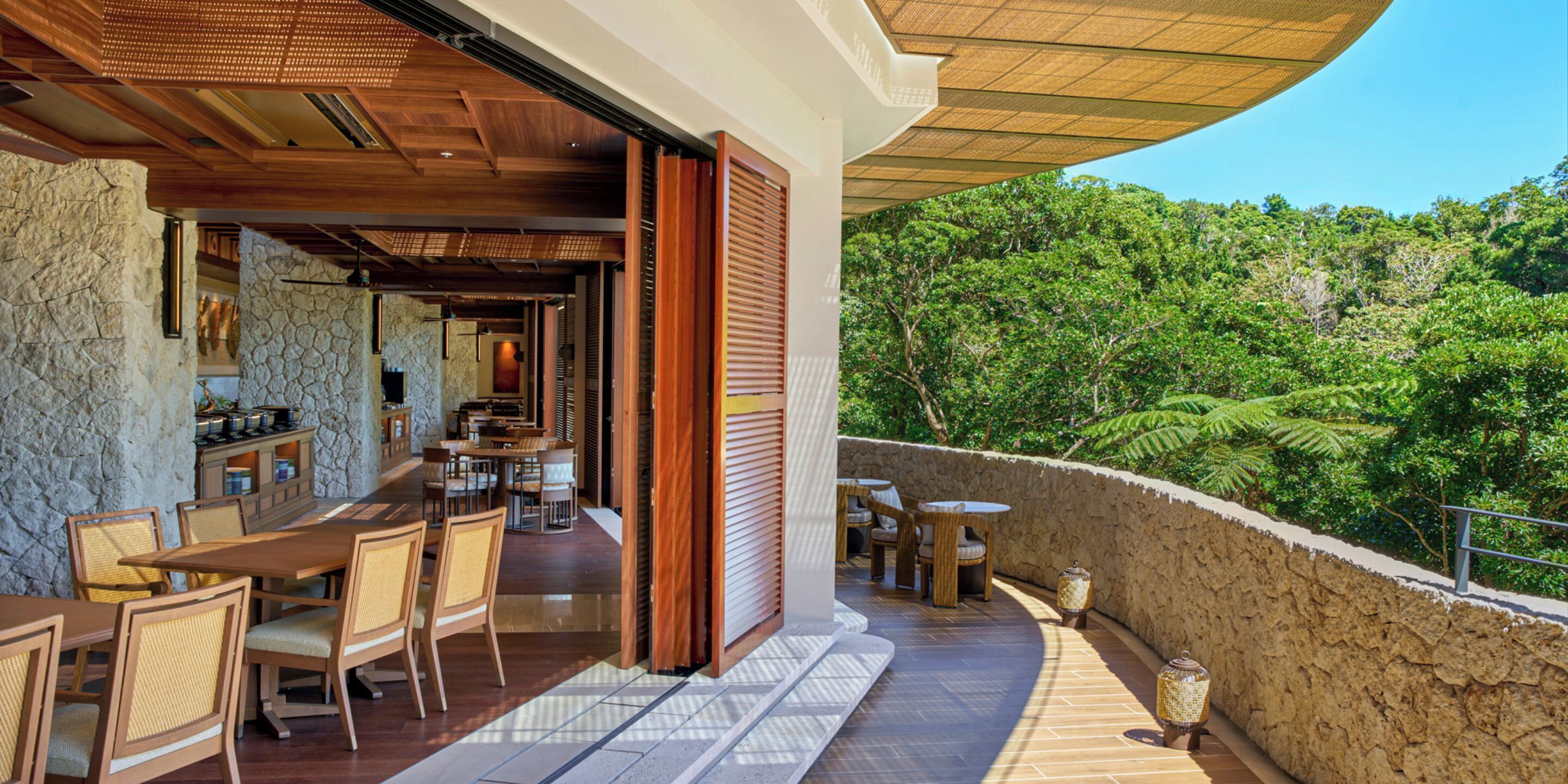 An outdoor dining area with tables and chairs, a stone wall, and a view of a forested landscape.
