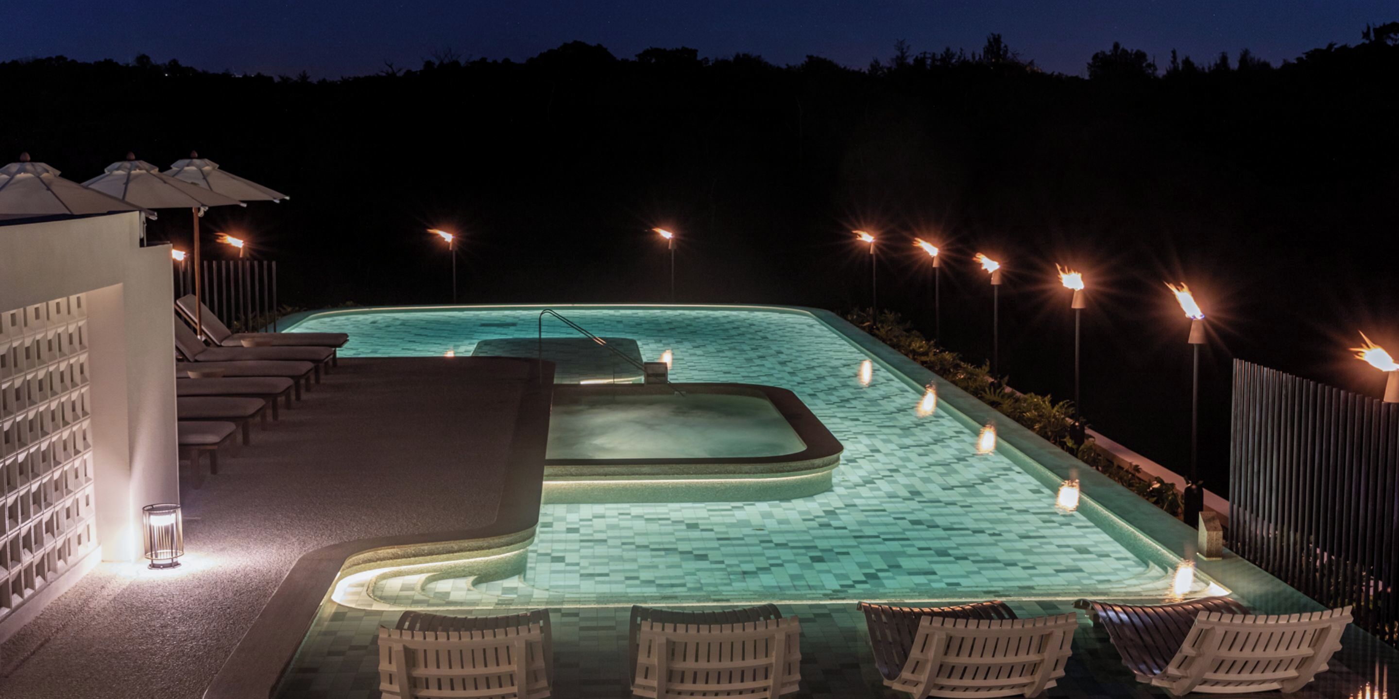 Una piscina iluminada de noche con sillones de descanso y antorchas.
