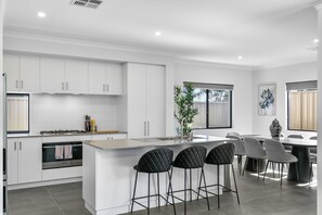 A modern kitchen with white cabinetry, a central island with bar stools, a dining area with a round table, and a large window providing natural light. - Bloom Brick A Sepal Stay (Perth)