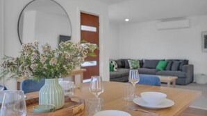 Ein Essbereich mit Holztisch, gedeckt für eine Mahlzeit, eine Vase mit Blumen und im Hintergrund ein Wohnzimmer mit Sofa und Couchtisch.