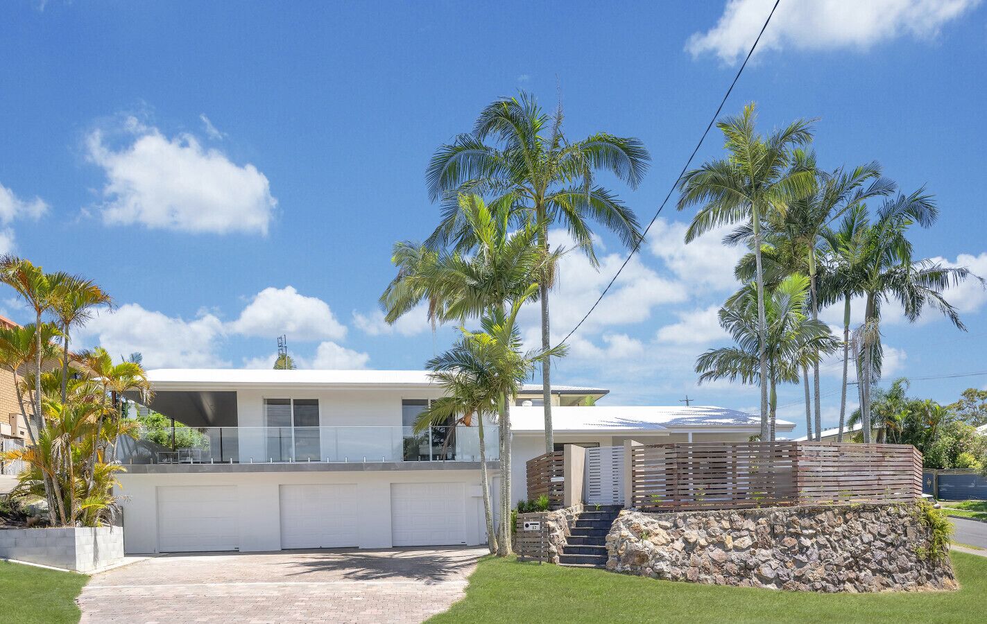 A modern two-story house with a garage, a stone retaining wall, and palm trees in the yard.