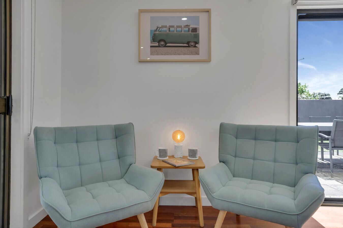 A room with two light blue armchairs, a small wooden table with a lamp, and a framed picture on the wall.