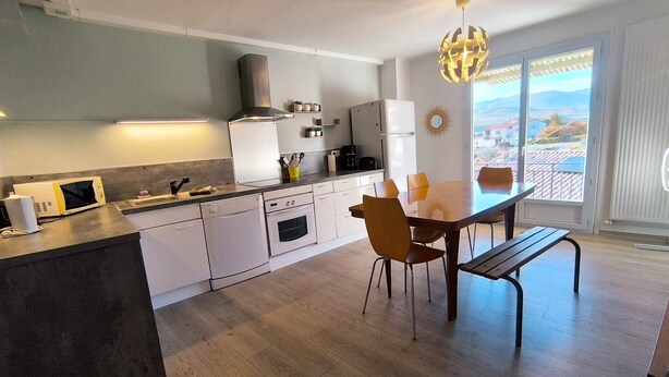 A modern kitchen with white cabinetry, a dining area with a wooden table and chairs, and a large window offering a view of the landscape. - La Maison de la Baume (Volonne)