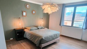 A bedroom with a bed, bedside lamps, a wall-mounted light fixture, and a window with a view of buildings. - La Maison de la Baume (Volonne)